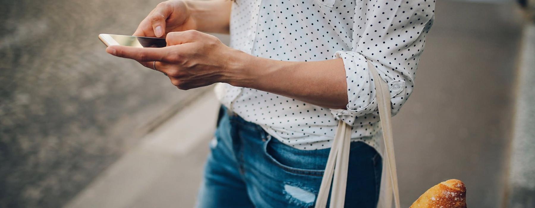 woman walks down the street and texts with a bag of groceries on her arm