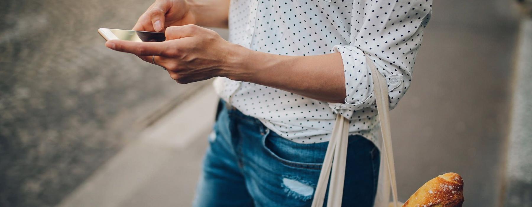 woman walks down the street and texts with a bag of groceries on her arm